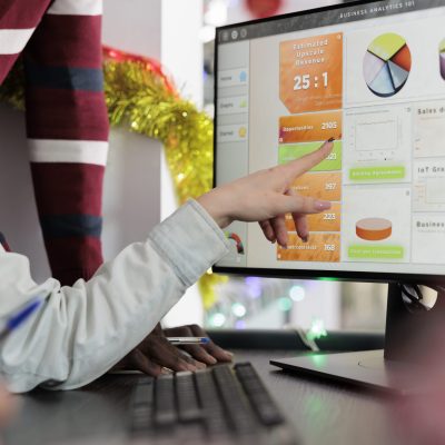 Businesswoman in Christmas decorated office showing coworkers key data point on computer screen needed for marketing insight. Employees working together on finishing project before impending deadline